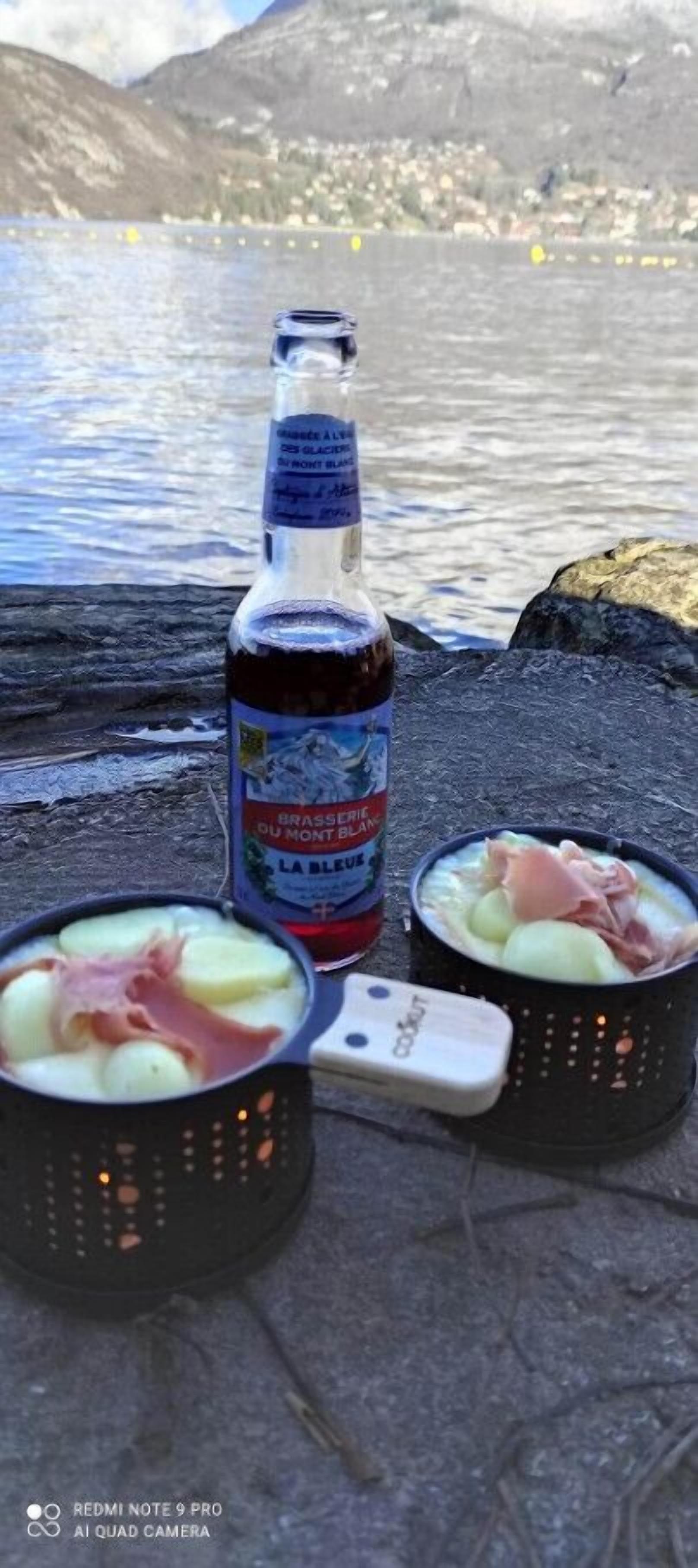 Una botella de cerveza Brasserie du Mont Blanc, dos tazones de sopa con jamón y un teléfono inteligente sobre una superficie de piedra junto a un cuerpo de agua.