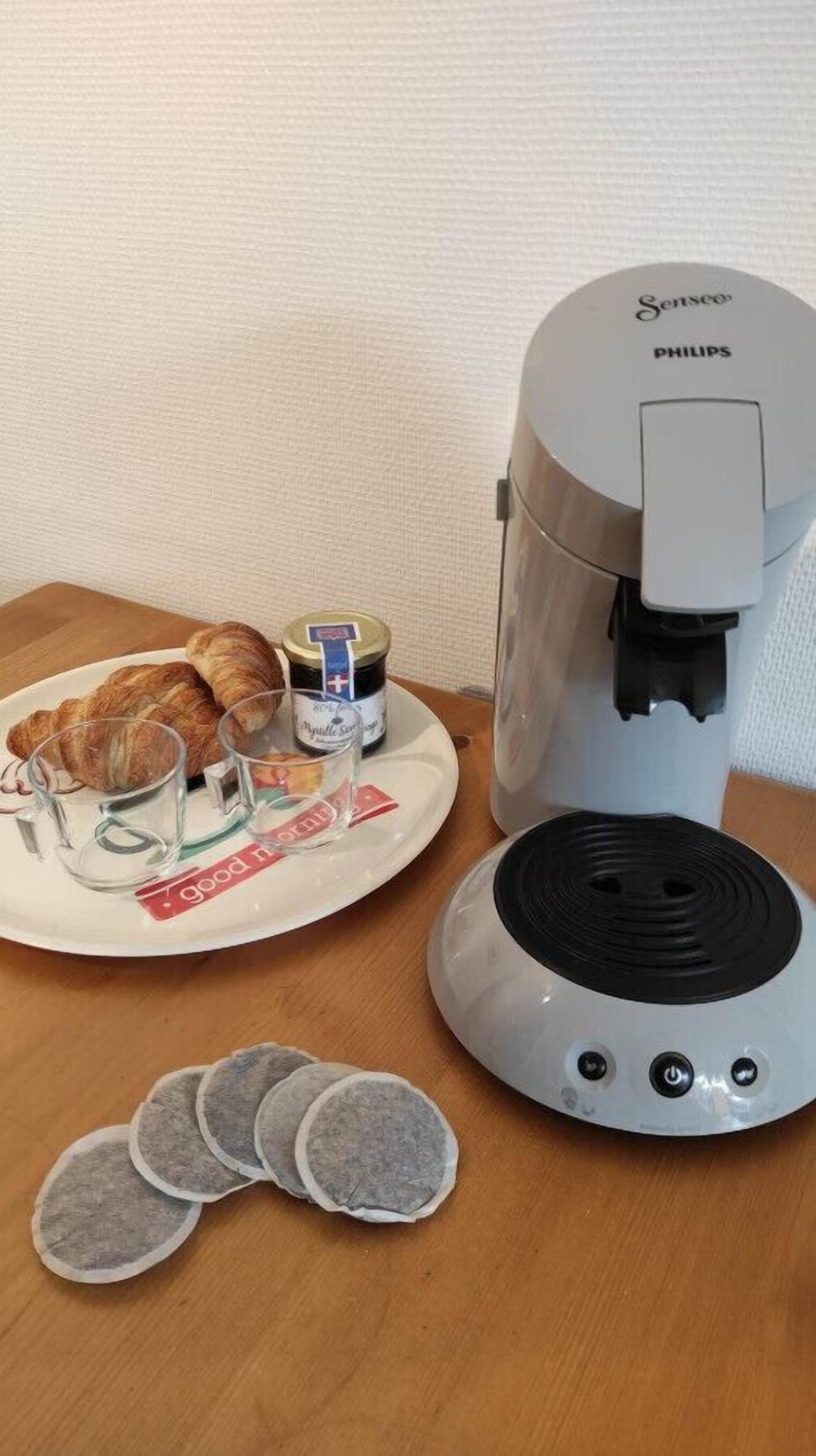 Een koffiezetapparaat, croissants, een pot jam en een bord met glazen op een houten tafel.