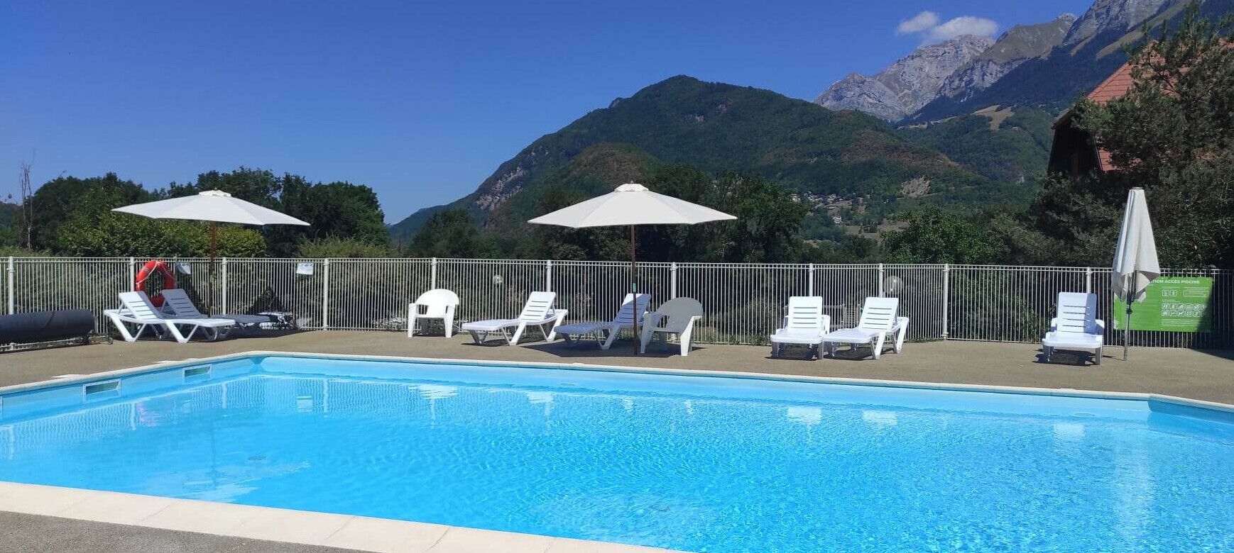 En swimmingpool med vita solstolar och parasoller, omgiven av ett metallstaket, med berg i bakgrunden.