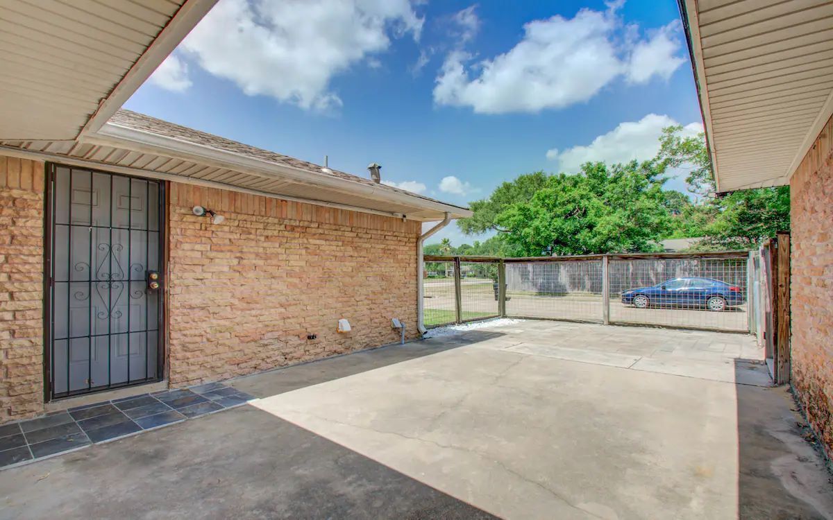 A brick house with a covered patio, a metal gate, and a blue car in the background.
