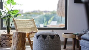 Une chaise en osier, un pouf rond, un panier tressé et un jardinier en bois près d’une fenêtre donnant sur un paysage verdoyant.