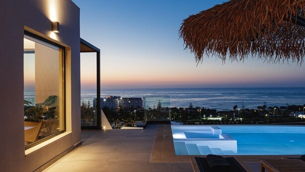 Un espace piscine extérieur moderne, avec vue sur l’océan au crépuscule, agrémenté d’un parasol en chaume et de mobilier de salon contemporain.