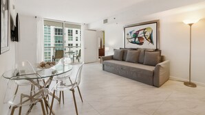A modern living room with a glass dining table, clear chairs, a grey sofa, and a colorful abstract painting.