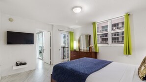 A bedroom with a bed, a TV mounted on the wall, a dresser, and a window with curtains.