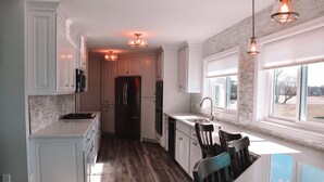 A modern kitchen with white cabinetry, a stainless steel refrigerator, a dining area with a blue table, and a window with a view of the outdoors. - Waterfront Paradise on the Eastern Shore of Chesapeake Bay  (Rock Hall)