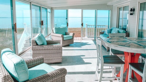 A sunlit outdoor patio with wicker furniture, a round table, and a view of the beach. - Waterfront Paradise on the Eastern Shore of Chesapeake Bay  (Rock Hall)