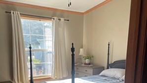 Un dormitorio con cama con dosel, una mesita de noche con lámpara, una ventana con cortinas y un ventilador de techo.