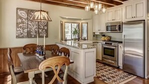 A kitchen with white cabinets, stainless steel appliances, a wooden dining table, and a hanging light fixture. - Lake Home in the Pines with a Dodger history (Lake Arrowhead)