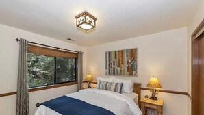 A bedroom with a large bed, two bedside lamps, a window with curtains, a ceiling light fixture, and a wall-mounted artwork. - Lake Home in the Pines with a Dodger history (Lake Arrowhead)