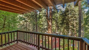 A wooden deck with a railing overlooking a forested area with tall trees and a red building in the background. - Lake Home in the Pines with a Dodger history (Lake Arrowhead)