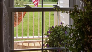A window with a view of a green landscape, an American flag, and a balcony with a railing. - 1880 Farmhouse With Direct Access to Tall Pines ATV Park (Andover)