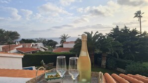 Ein Tablett mit einer Champagnerflasche, zwei Gläsern und einer kleinen Oliven-Schale auf einem Balkon mit Blick auf eine Küstenlandschaft, Häuser und Palmen.