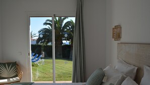 Ein Schlafzimmer mit Bett, Kissen und Blick auf eine Rasenfläche sowie Palmen durch eine Schiebetür, inklusive einer Wandleuchte.