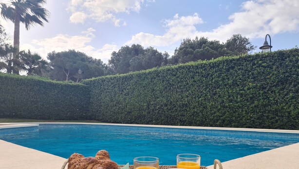Ein Frühstücksbrettchen am Pool mit Croissants, Orangensaft und Kaffee, mit Blick auf ein klares, blaues Schwimmbecken mit einer Hecke und einer Lampe im Hintergrund.