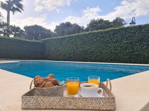 Bandeja de desayuno junto a la piscina, con croissants, jugo de naranja y café, con vistas a una piscina azul cristalina y un seto con una lámpara al fondo.