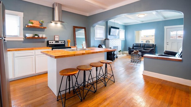 Una cocina moderna con una isla central, taburetes de bar y una zona de comedor con sofá y televisor.