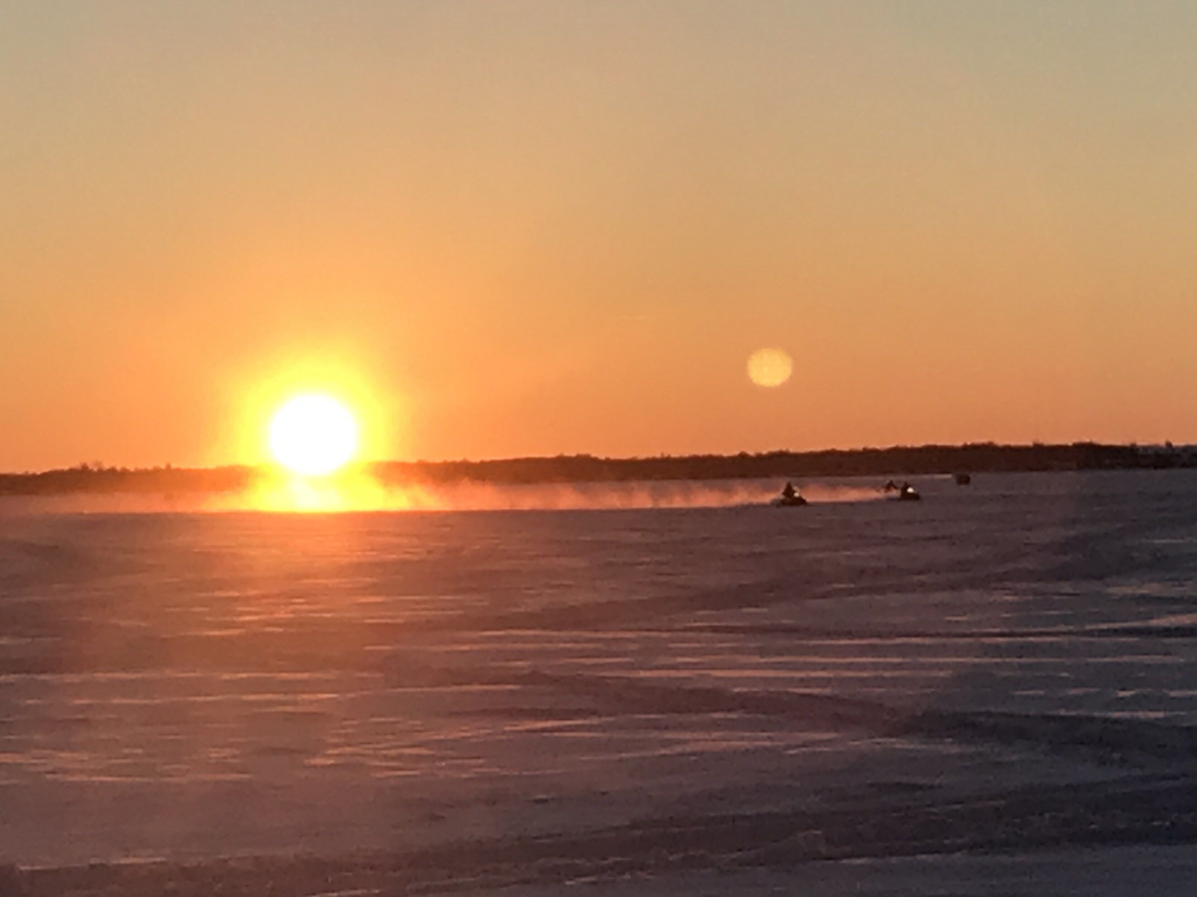 Un coucher de soleil sur une étendue d’eau, avec une côte lointaine et quelques bateaux.