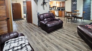 Un vaste salon avec un canapé en cuir, un coin repas avec une table et des chaises, une cuisine à l’arrière-plan et un ventilateur de plafond.