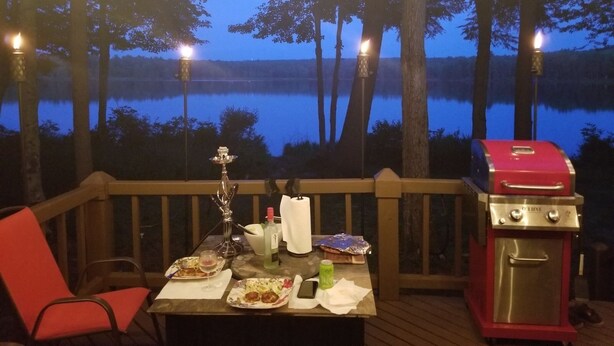 Une table à manger avec des assiettes remplies de nourriture, un barbecue et une vue sur un lac entouré d’arbres.