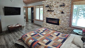 Une chambre avec un plafond en bois, un ventilateur de plafond, un lit recouvert d’une courtepointe, un mur en pierre, une cheminée et un téléviseur fixé au mur.