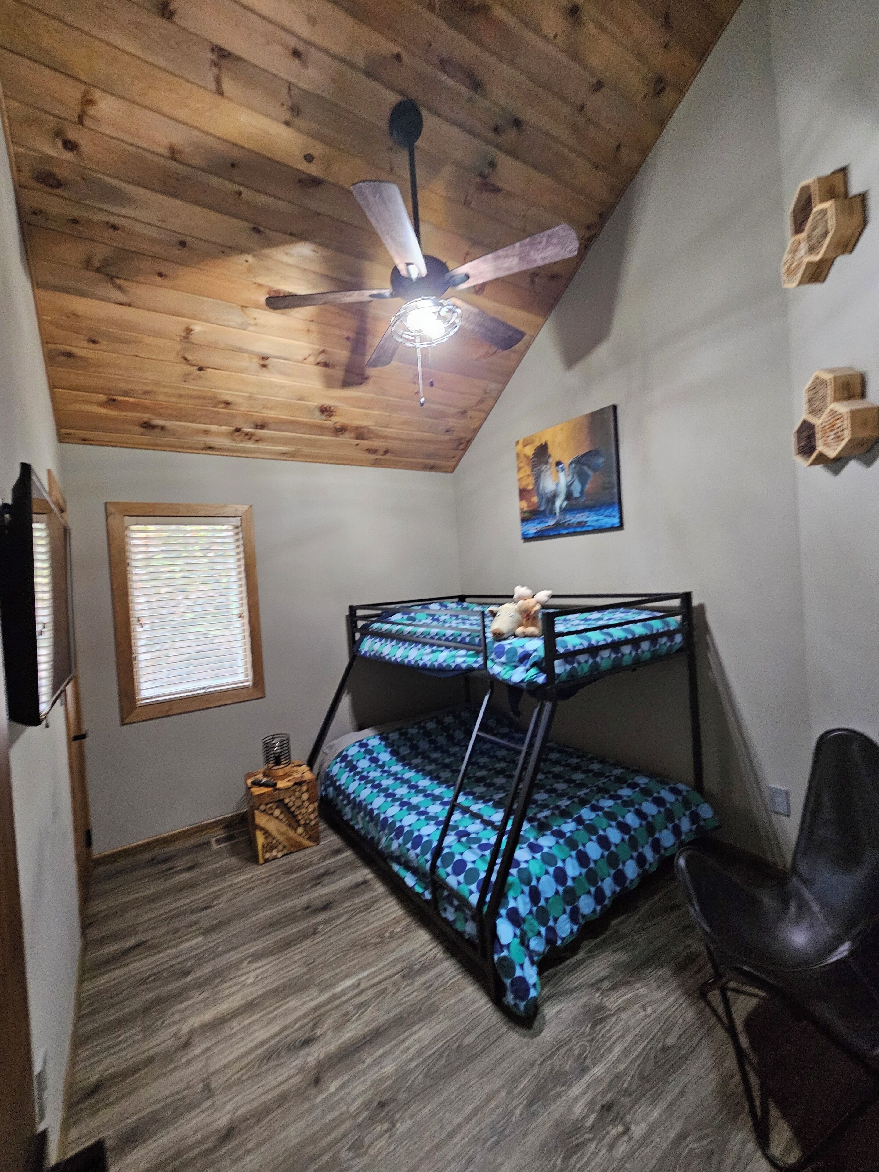 A bunk bed room with a ceiling fan, wooden walls, a painting, and a window with blinds.