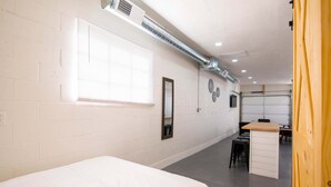 A hotel room with a bed, a desk, and a chair. There is a window with blinds, a mirror, and a wall-mounted air vent. - Quaint cozy studio located in the heart of El Paso  (El Paso)