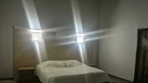Een kleine, zwak verlichte kamer met een enkel bed, een klein tafeltje en twee wandlampen.