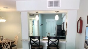A living room with a beige sofa and patterned throw pillow, a dining area with wooden chairs and a table, and a kitchen area with a refrigerator and a sink. - Fully remodeled, block to beach. 2-story, first floor entry, park at front door (Hilton Head Island)