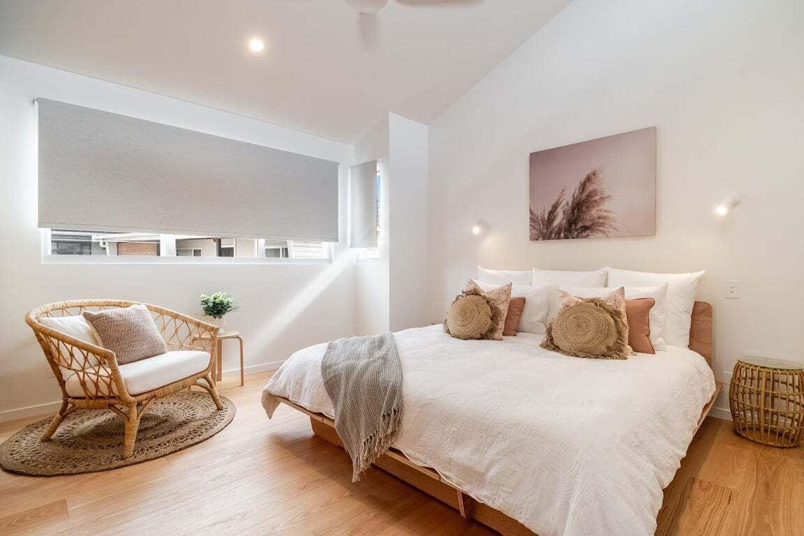 Une chambre à coucher avec un grand lit, une chaise en osier, une petite table avec une plante et une fenêtre avec des stores.