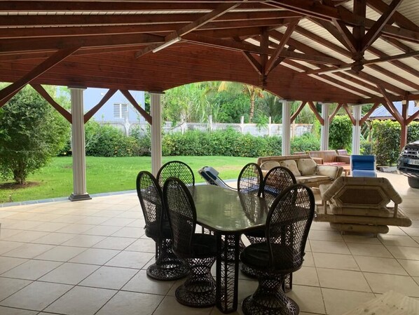 A covered patio area with a table and chairs, a barbecue grill, and a view of a garden and house in the background.