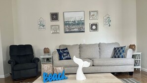 Un salon avec un canapé gris, un fauteuil noir et une table basse décorée d’une queue de baleine et portant l’inscription « Beach ».