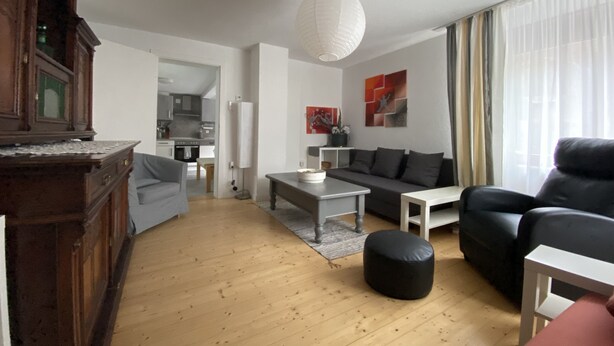 A modern living room with a wooden floor, a grey sofa, a black leather armchair, a white coffee table, and a dining area visible through an open door. - Casa Siepelunga (Villmar)