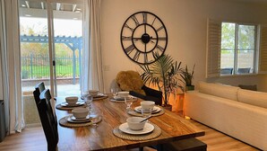 A dining area with a wooden table set for four, a large wall clock, and a view of the outdoors through a glass door. - Golf course house with 2 master bedrooms (Fontana)