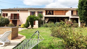 A two-story house with a tiled roof, a balcony, and a garden area with a hammock and a white chair. - Pleasures and Idleness... (Boisredon)