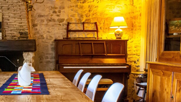 A wooden dining table with white chairs, a piano, and a lamp in a room with stone walls. - Pleasures and Idleness... (Boisredon)