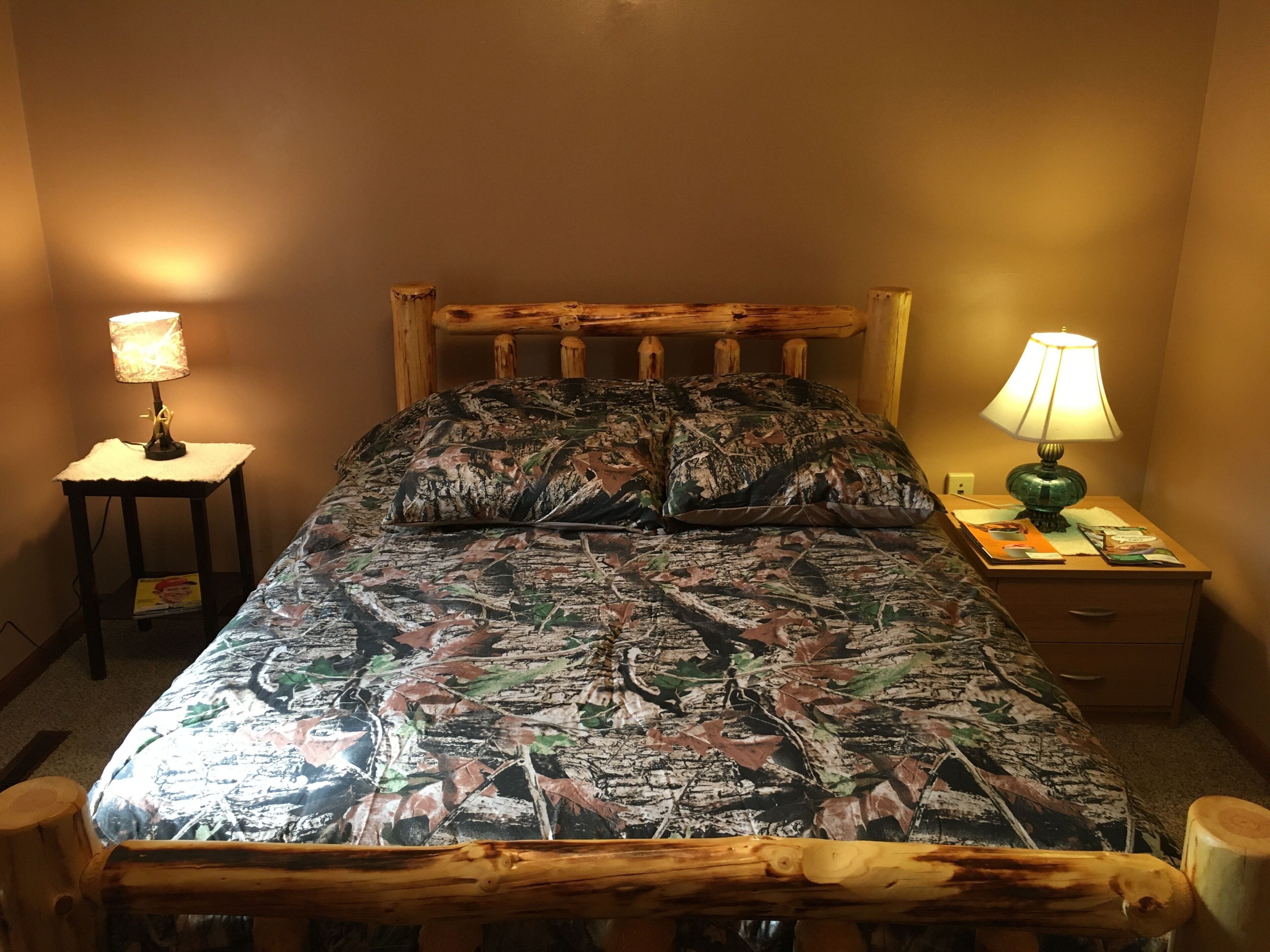 A bed with a camouflage-patterned bedspread, two bedside lamps, a wooden headboard, and a small table with a lamp and a book.
