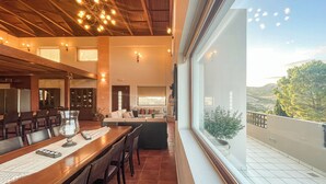 A modern dining area with a long wooden table, multiple chairs, and a large window offering a view of the outdoors. - Luxurious Panoramic View Villa (Archanes-Asterousia)