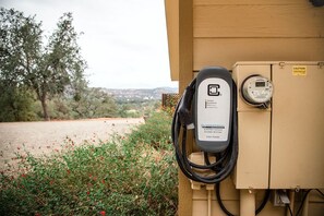 An electric vehicle charging station attached to a utility box with a digital display, set against a backdrop of trees and a dirt path. - Sierra Ranch - Yosemite Views, Cozy Fireplaces & Stunning Sunsets (Mariposa)