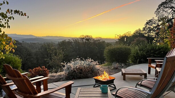 Une terrasse avec des meubles d’extérieur, un foyer et une vue panoramique sur les montagnes au coucher du soleil.