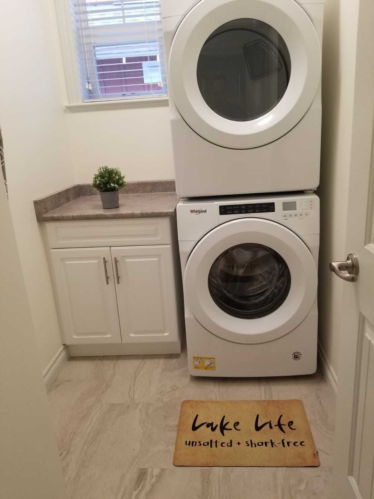 Une buanderie équipée de deux machines à laver et deux sèche-linge superposés, d’une petite plante en pot posée sur le comptoir et d’un tapis sur lequel est inscrit « Lake Life ».