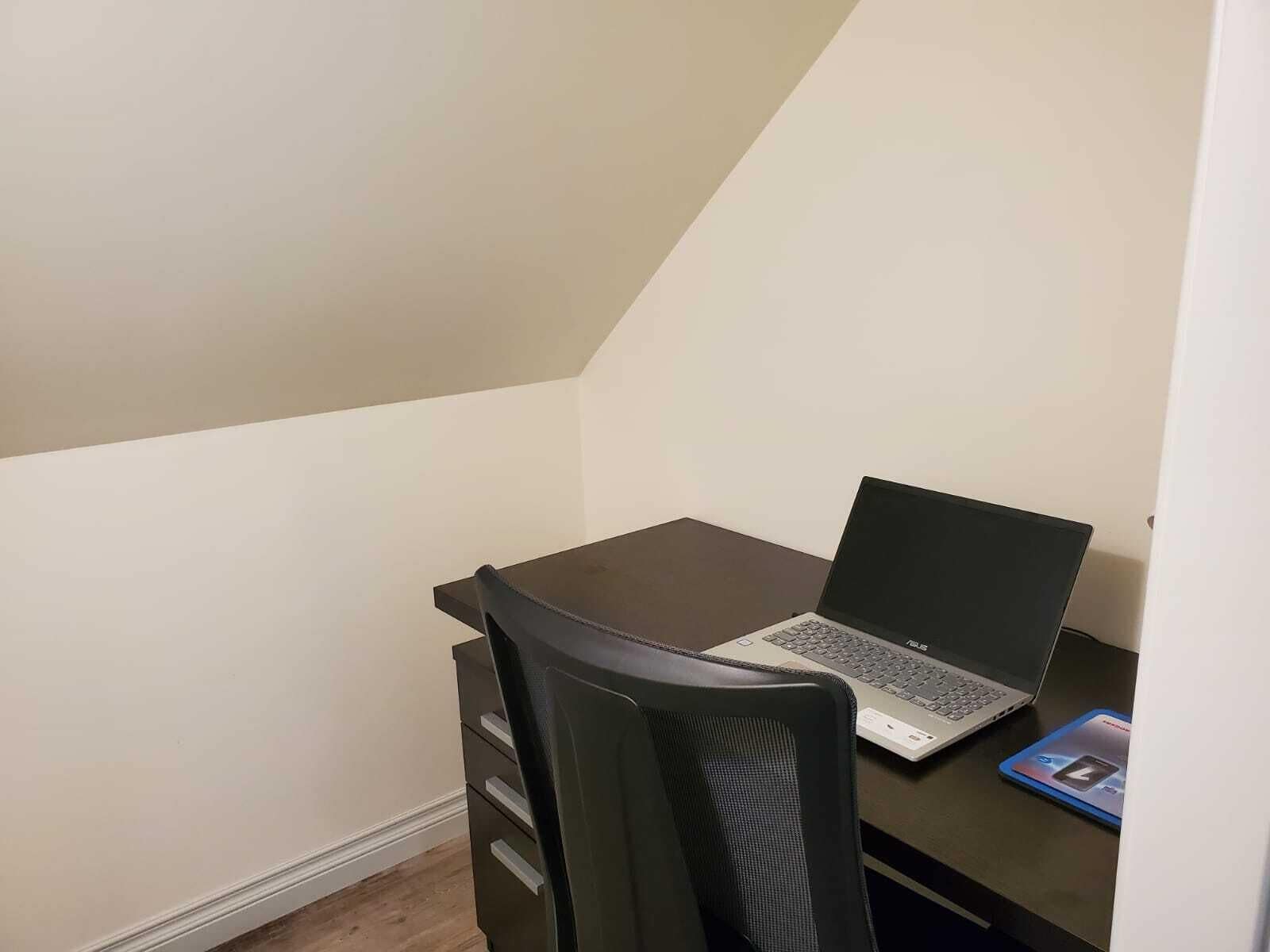 Un escritorio de madera oscuro y pequeño con una computadora portátil y una tableta en un rincón de una habitación con techos inclinados y paredes lisas.