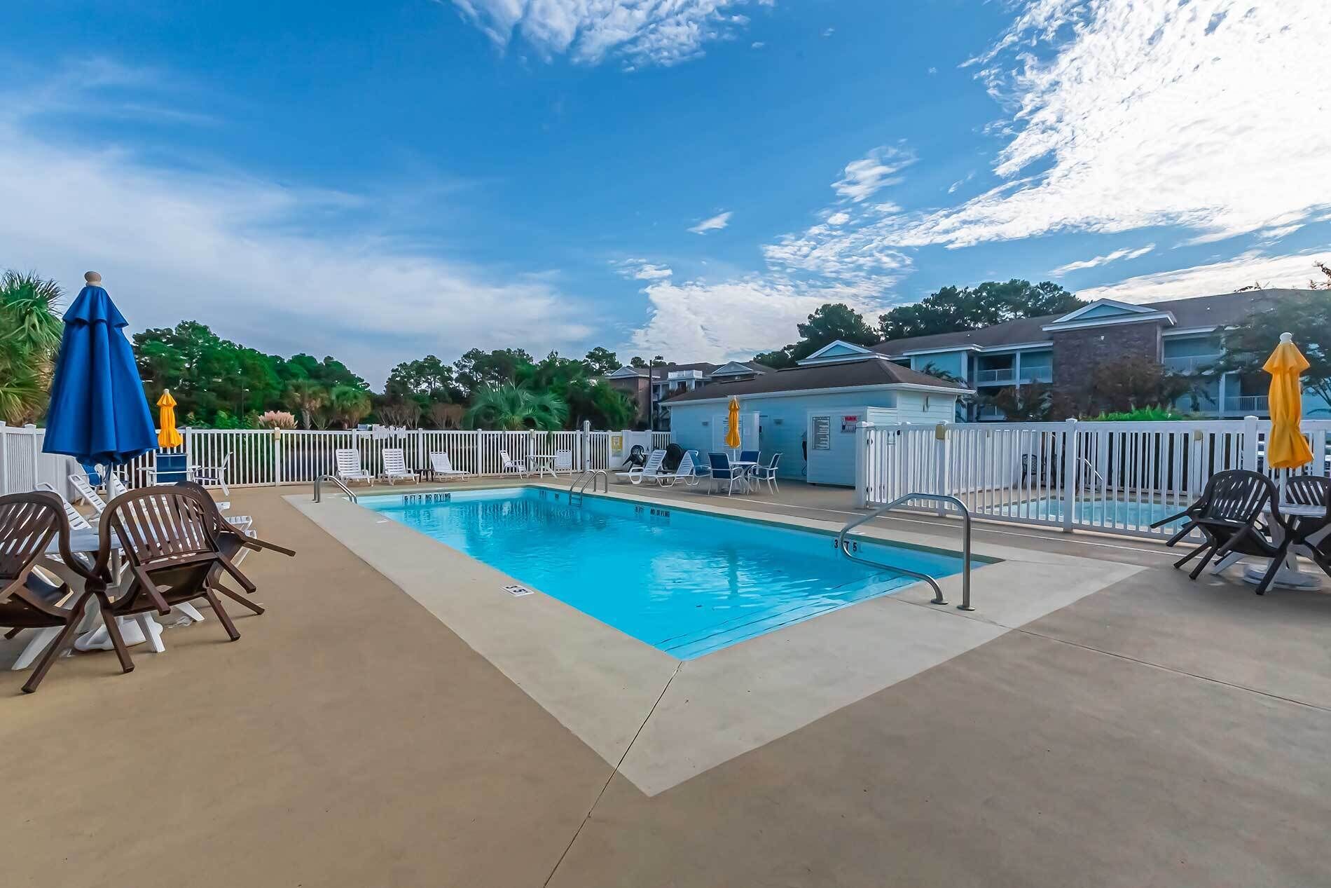 A swimming pool with lounge chairs and umbrellas, surrounded by a white fence and residential buildings.