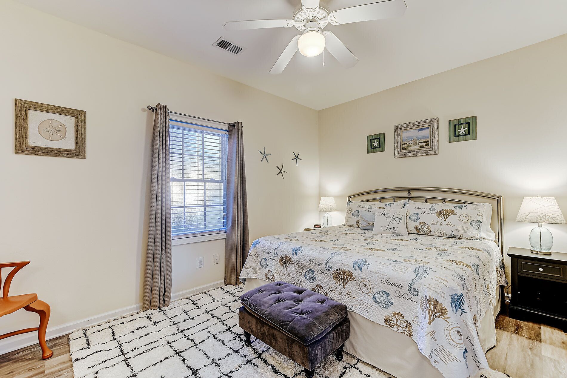 A bedroom with a bed, a nightstand, a lamp, a chair, a window with curtains, a ceiling fan, and wall decorations.