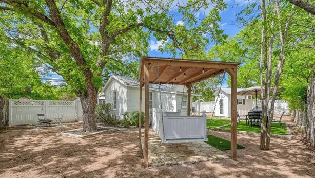 A covered outdoor seating area with a wooden pergola, stone pathway, and a white fence in the background. - Romantic-Steps To Main-FirePit! (Fredericksburg)