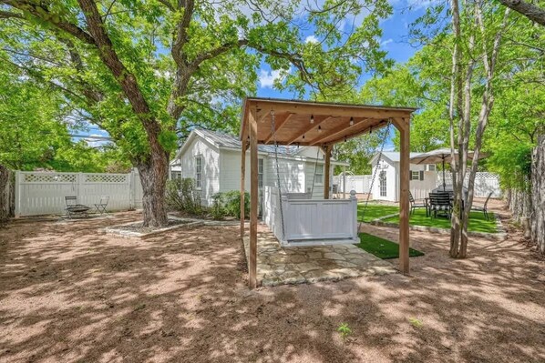 A covered outdoor seating area with a wooden pergola, stone pathway, and a white fence in the background.