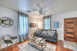 Ein Wohnzimmer mit grauem Sofa, einem Glastisch, einem grauen Sessel, einer Holztür und einem Deckenventilator.