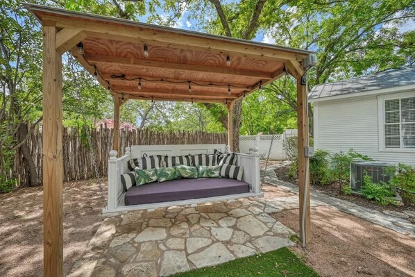 Una pérgola de madera con un asiento tipo columpio y cojines, rodeada por un patio de piedra y una valla de madera.