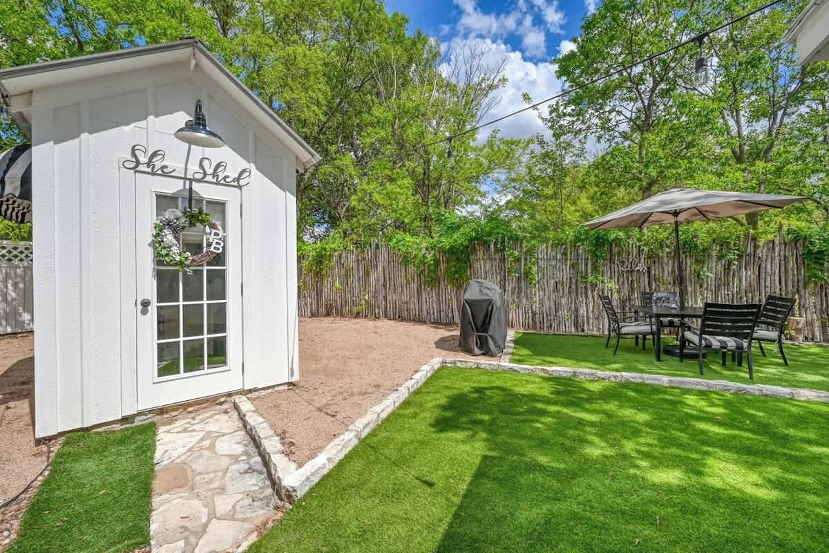 A white shed with a door and a window, a covered grill, a table with chairs, and a wooden fence.