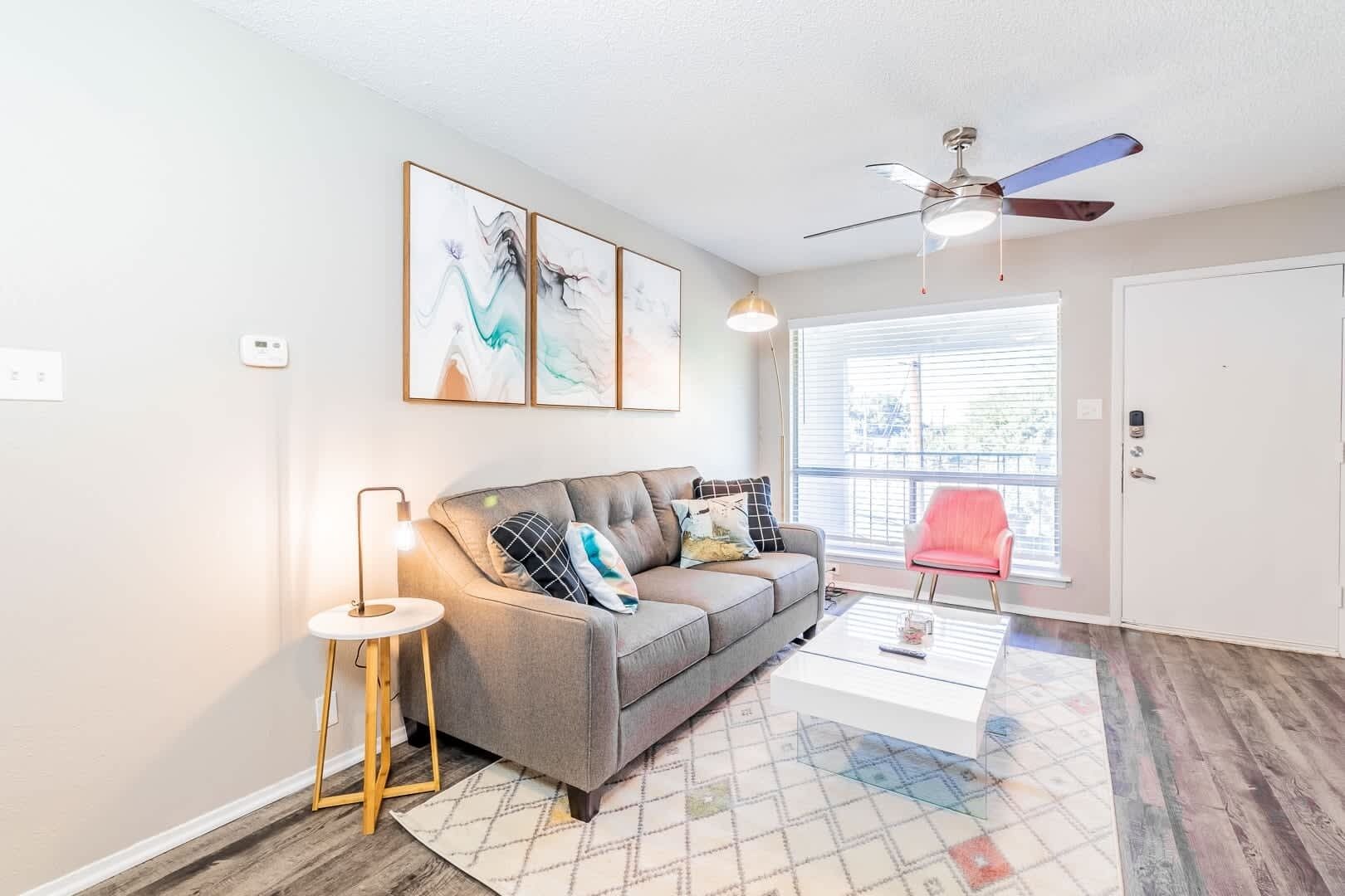 A living room with a grey sofa, a coffee table, a pink chair, a ceiling fan, and a window with blinds.