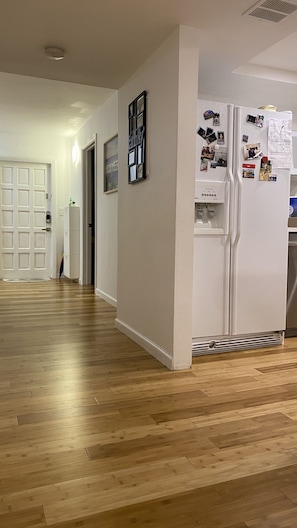 Enter a welcoming hallway with a wooden floor, leading to a white door and a cozy space with a refrigerator and personal touches.